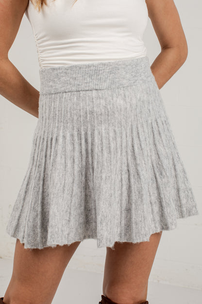 A light gray ribbed knit pleated mini skirt worn by a model, paired with a white tank top against a white wall.