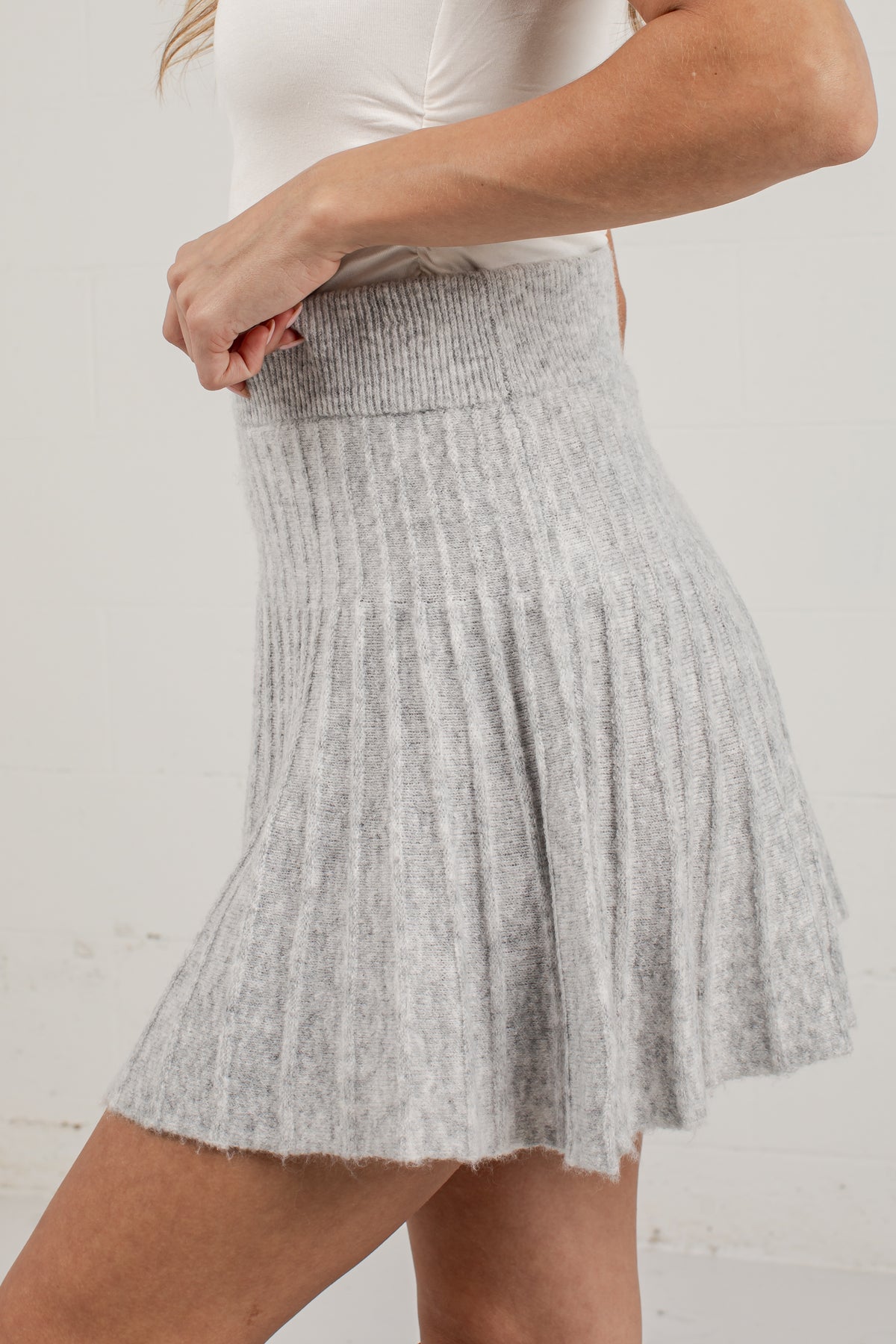 A light gray ribbed knit pleated mini skirt worn by a model, paired with a white top, against a white brick wall background.