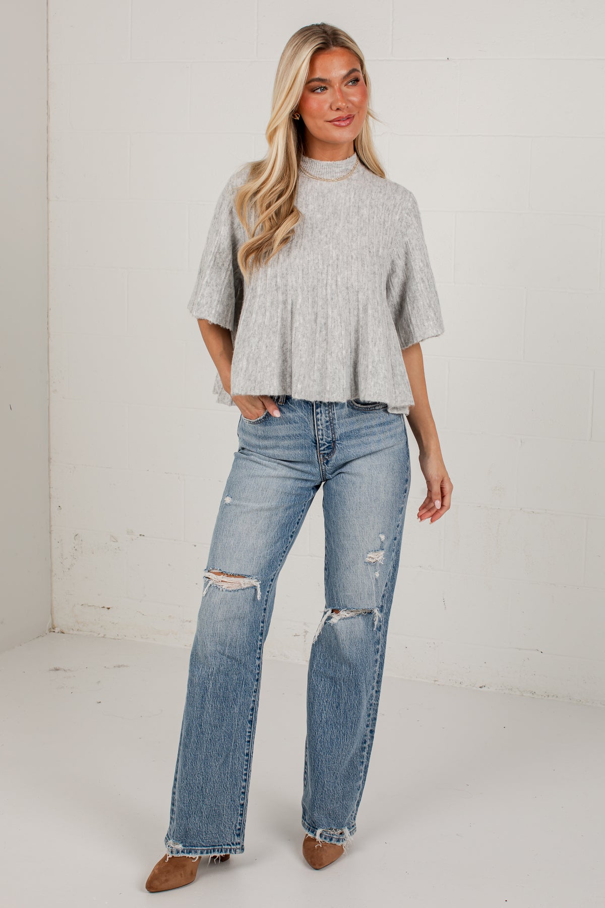 A woman modeling a light gray short-sleeve mock neck knit top and light-wash distressed blue jeans, standing against a white studio wall.