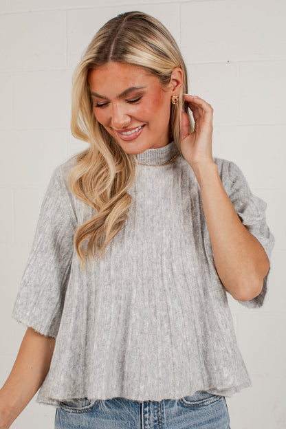 A woman wearing a light gray heathered short-sleeve mock neck top with a textured crinkle fabric, paired with blue jeans, standing against a white brick wall.