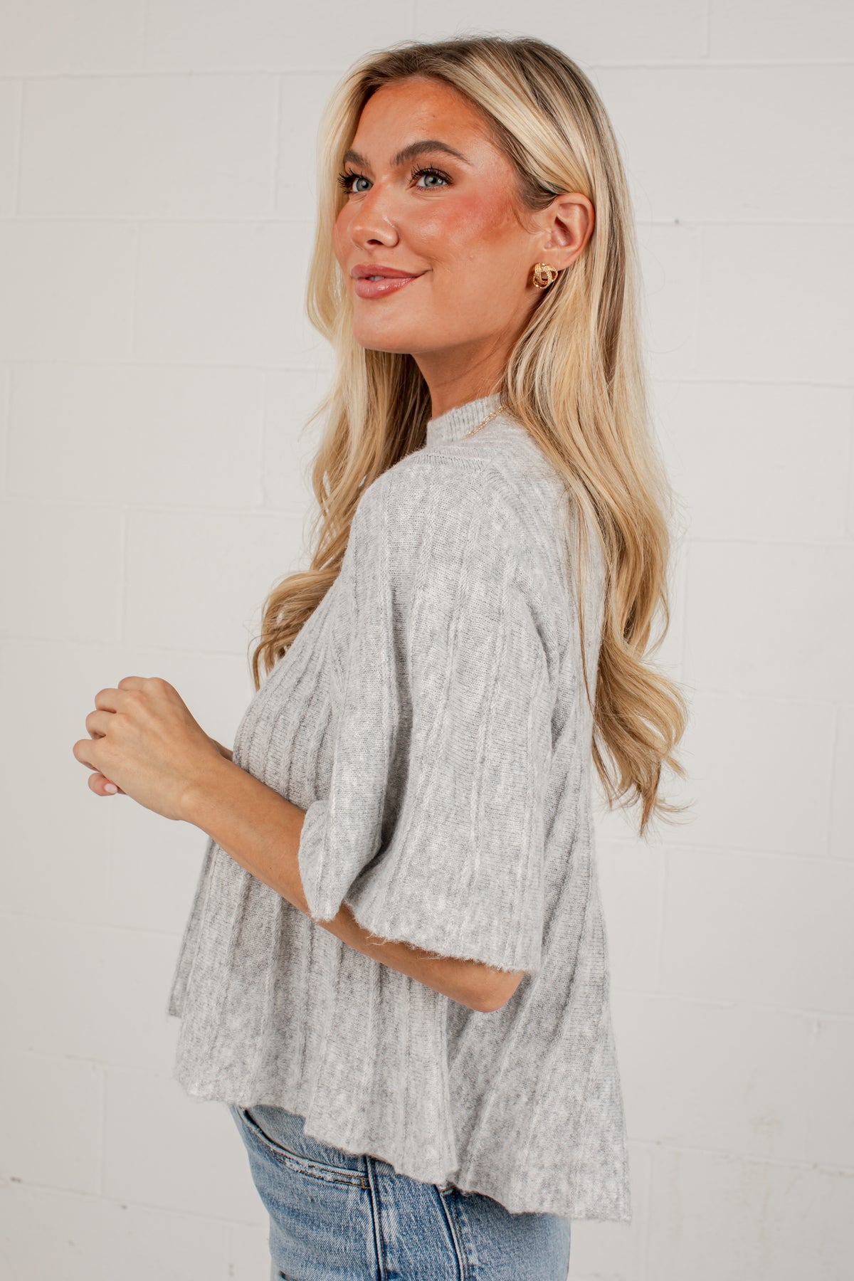 A woman with blonde hair wearing a light gray heathered knit top with three-quarter sleeves and blue jeans, posing against a white brick wall.