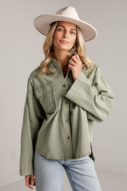 A woman wearing an oversized sage green button-up shirt with chest pockets, light-wash jeans, and a wide-brimmed cream hat, standing against a light gray wall.
