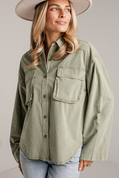 A woman wearing an oversized sage green button-up shirt with chest pockets, paired with light blue jeans and a wide-brimmed hat, photographed against a light gray wall.