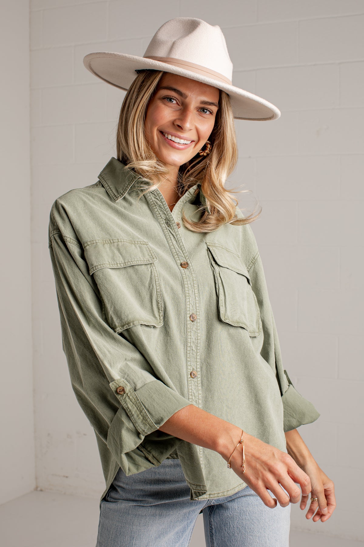 A woman wearing a light sage green button-up shirt with chest pockets, light-wash jeans, and a wide-brimmed cream hat, standing against a white paneled wall.