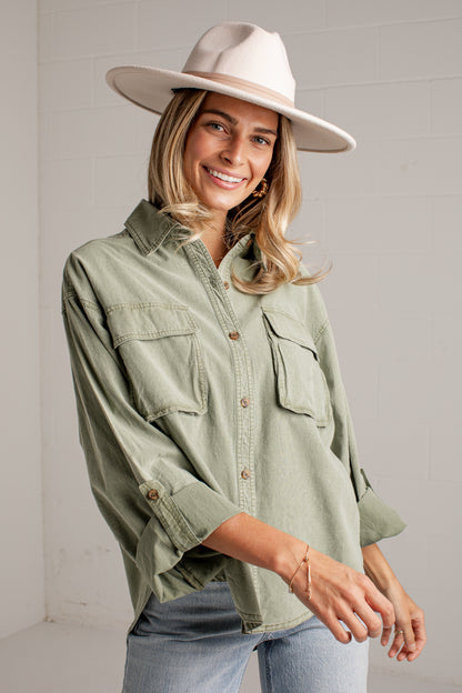 A woman wearing a light sage green button-up shirt with chest pockets, light-wash jeans, and a wide-brimmed cream hat, standing against a white paneled wall.