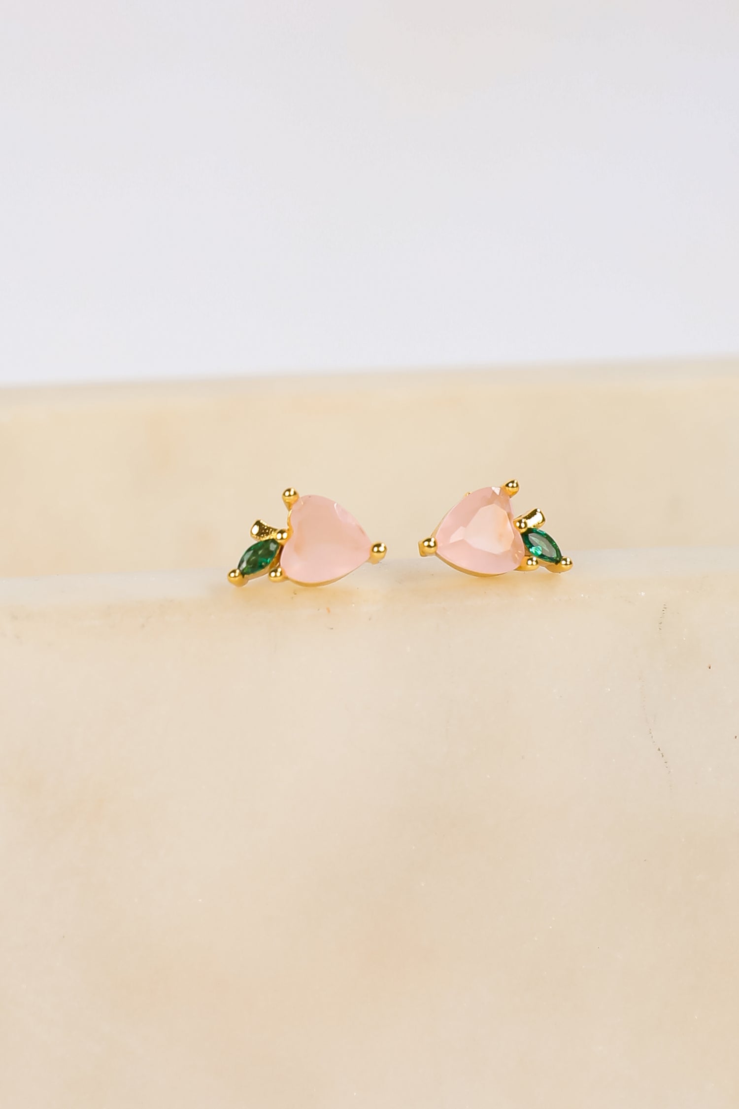 Pair of pink heart-shaped earrings with green gemstones on a beige surface.