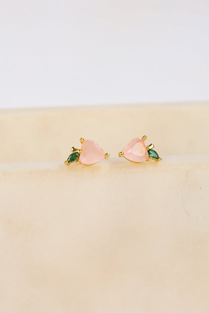 Pair of pink heart-shaped earrings with green gemstones on a beige surface.