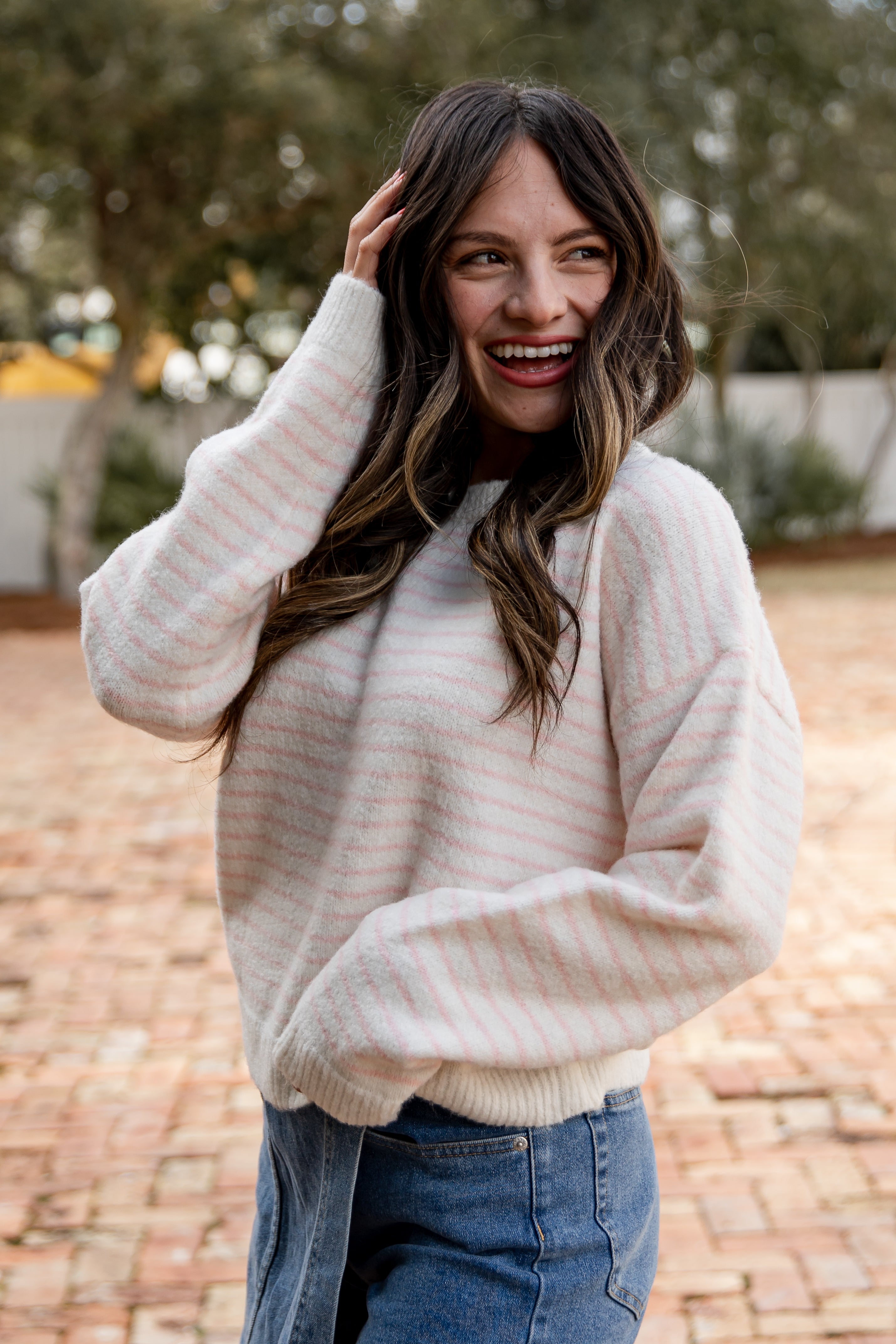 Woman wearing a cream sweater and blue jeans outdoors on a brick path.
