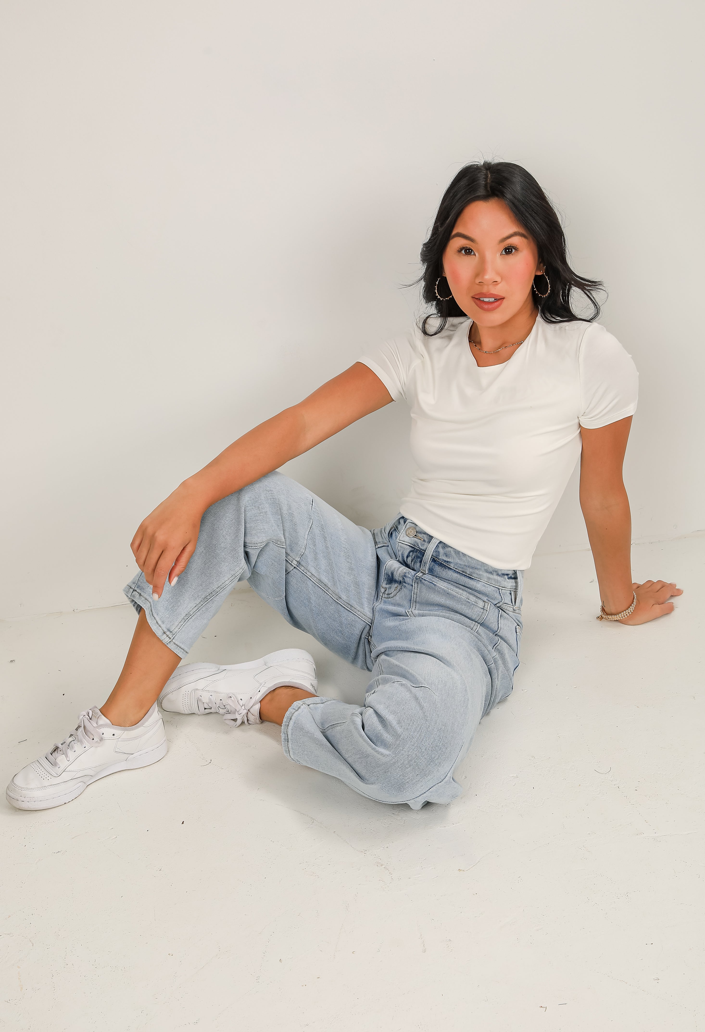 Woman sitting on a white floor wearing a white t-shirt and light blue jeans.