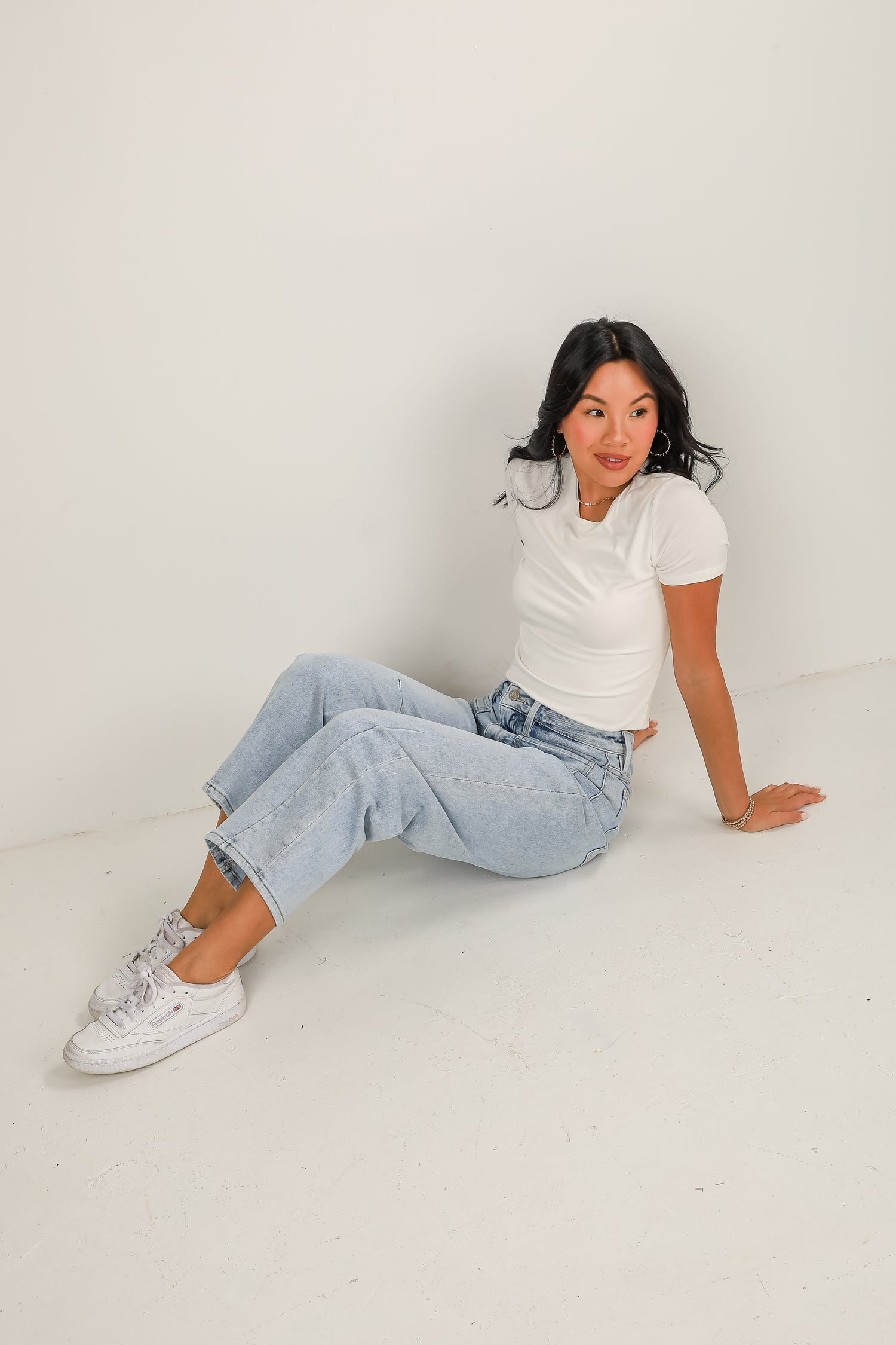 Woman sitting on a white floor wearing a white t-shirt, light blue jeans, and white sneakers.