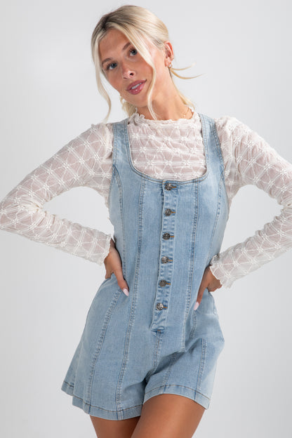 Woman wearing a denim overall dress with a white lace top on a plain background