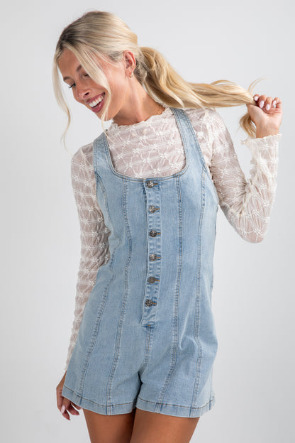 Woman wearing a denim overall romper with a white lace top on a plain background