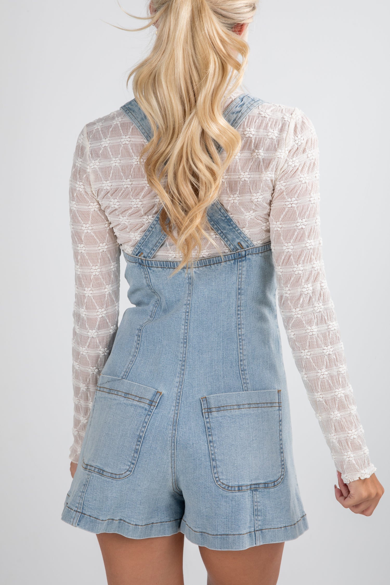 Person wearing a denim overall dress with a white lace top on a plain background