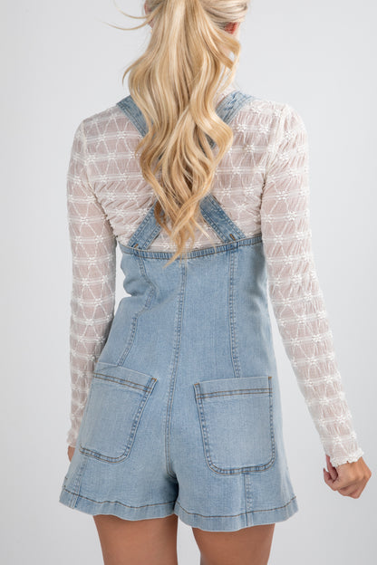 Person wearing a denim overall dress with a white lace top on a plain background