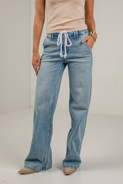 A model wearing light-wash blue wide-leg jeans with a white drawstring waist, paired with a beige top and beige pointed-toe flats, standing against a white studio wall.