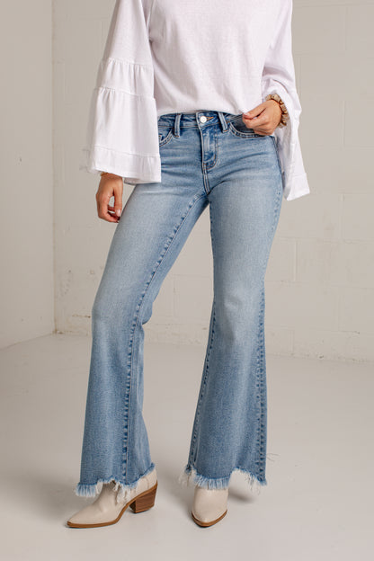 A model wearing light-wash blue flare jeans with frayed hems, paired with a white long-sleeve blouse and white heeled boots, standing against a light gray brick wall.