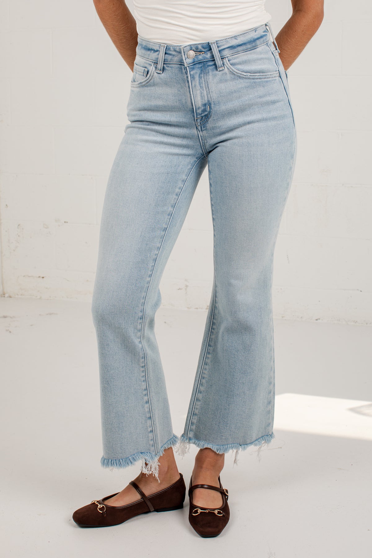 A person wearing light-wash blue flare jeans with a raw frayed hem, paired with a white top and dark brown flat shoes, standing against a white studio wall.