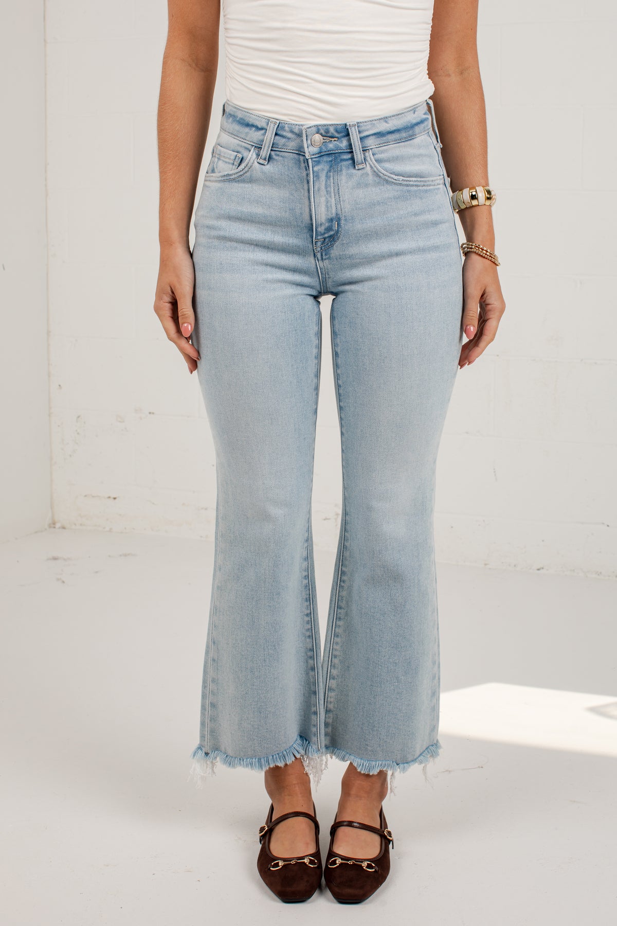 A model wearing light-wash blue flared jeans with a raw frayed hem, paired with a white top and dark brown flat shoes, standing against a white wall.
