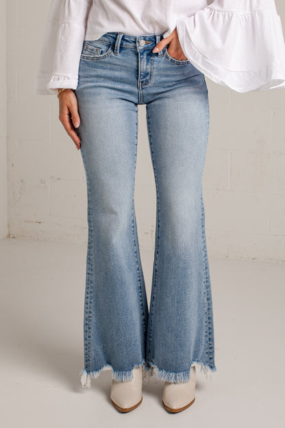 A person wearing light-wash blue flare jeans with a frayed hem, paired with a white blouse and white pointed-toe heels, standing against a light neutral wall.