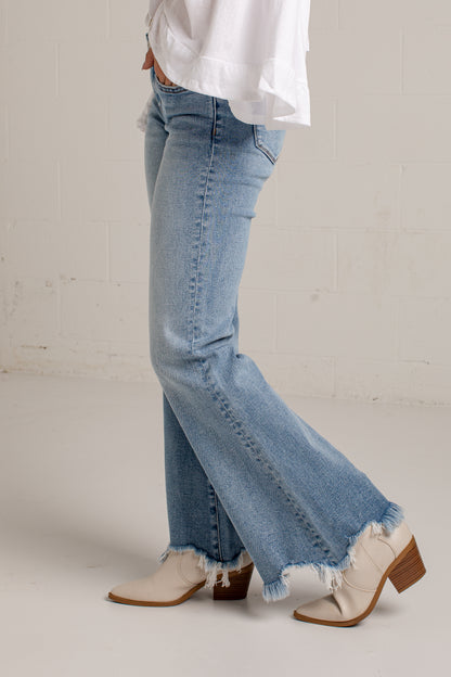 A person wearing light-wash blue flare jeans with a raw frayed hem, styled with a white top and beige heeled boots against a light neutral wall.