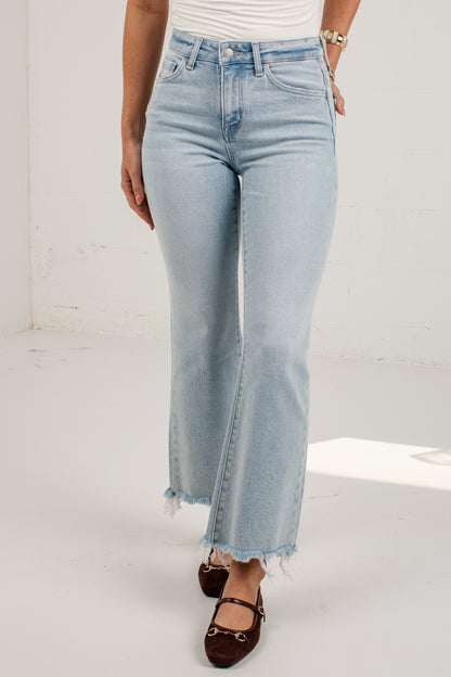 A model wearing light-wash blue flare jeans with a raw frayed hem, paired with dark brown flat shoes and a white top, standing against a white studio wall.