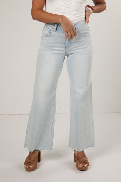 A model wearing light-wash, wide-leg denim jeans with a raw frayed hem, paired with a white top and brown heeled sandals, shown from the waist down against a plain studio background.
