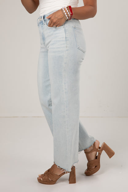 A person wearing light-wash, wide-leg cropped jeans with a frayed hem, paired with brown platform sandals and stacked pearl bracelets, shown from the waist down against a plain off-white studio background.