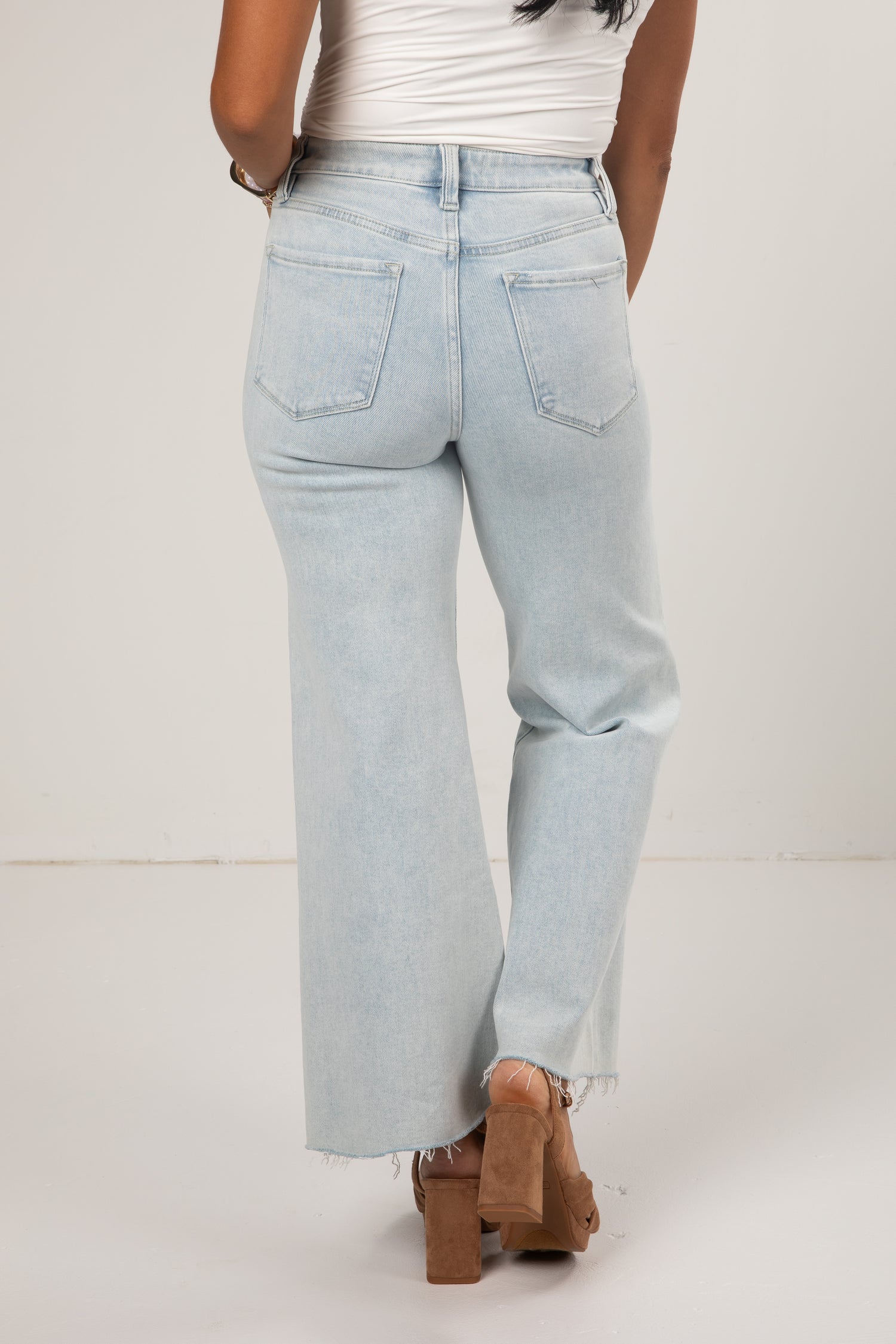 A person shown from the back wearing very light blue wide-leg jeans with a frayed hem, paired with a white top and brown block-heel sandals, against a plain off-white studio background.