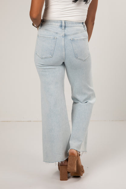 A person shown from the back wearing very light blue wide-leg jeans with a frayed hem, paired with a white top and brown block-heel sandals, against a plain off-white studio background.