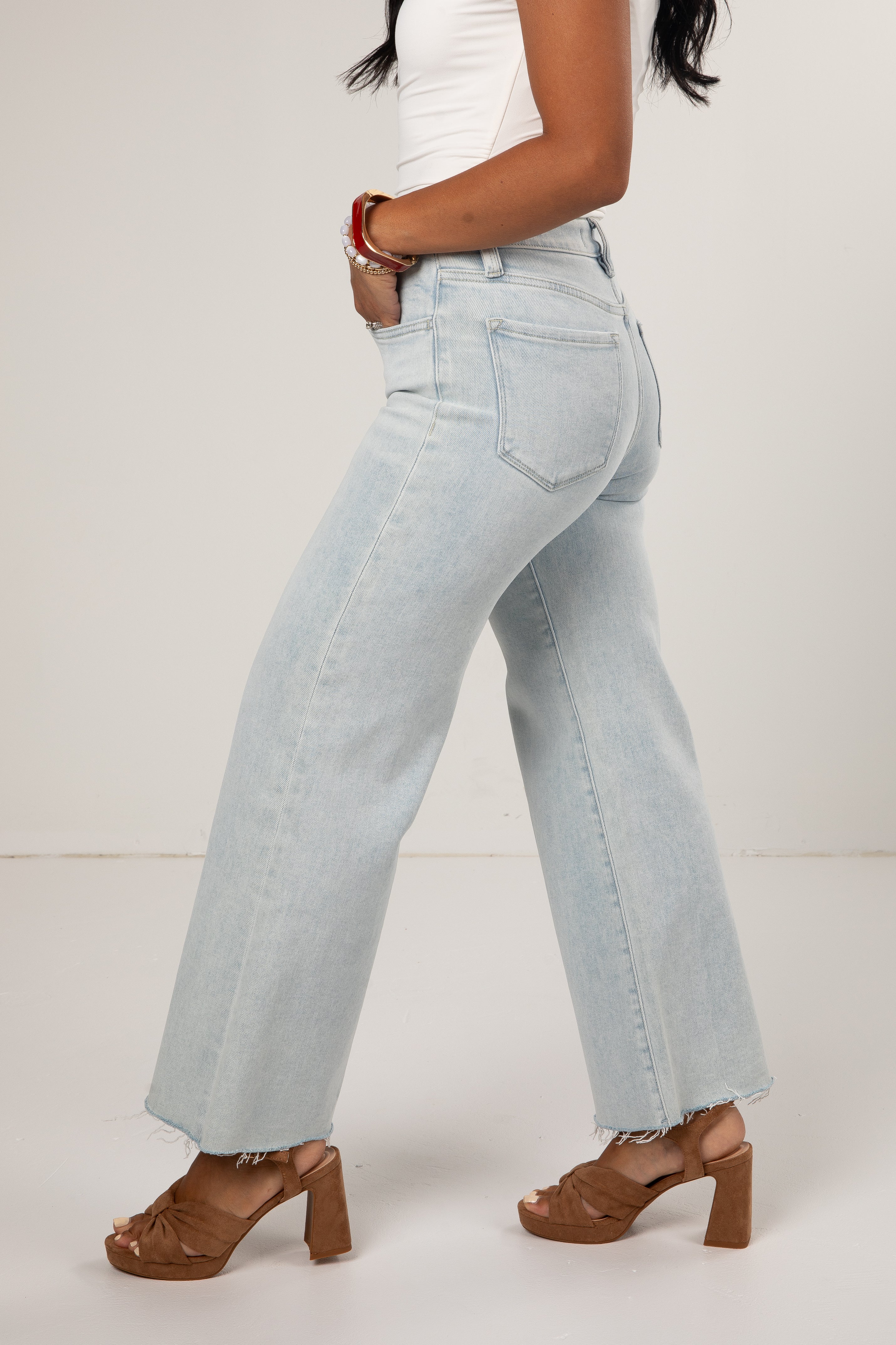 A side profile view of a model wearing light-wash, wide-leg denim jeans with a raw hem, styled with a white top and brown block-heel sandals against a plain off-white studio background.