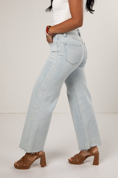 A side profile view of a model wearing light-wash, wide-leg denim jeans with a raw hem, styled with a white top and brown block-heel sandals against a plain off-white studio background.