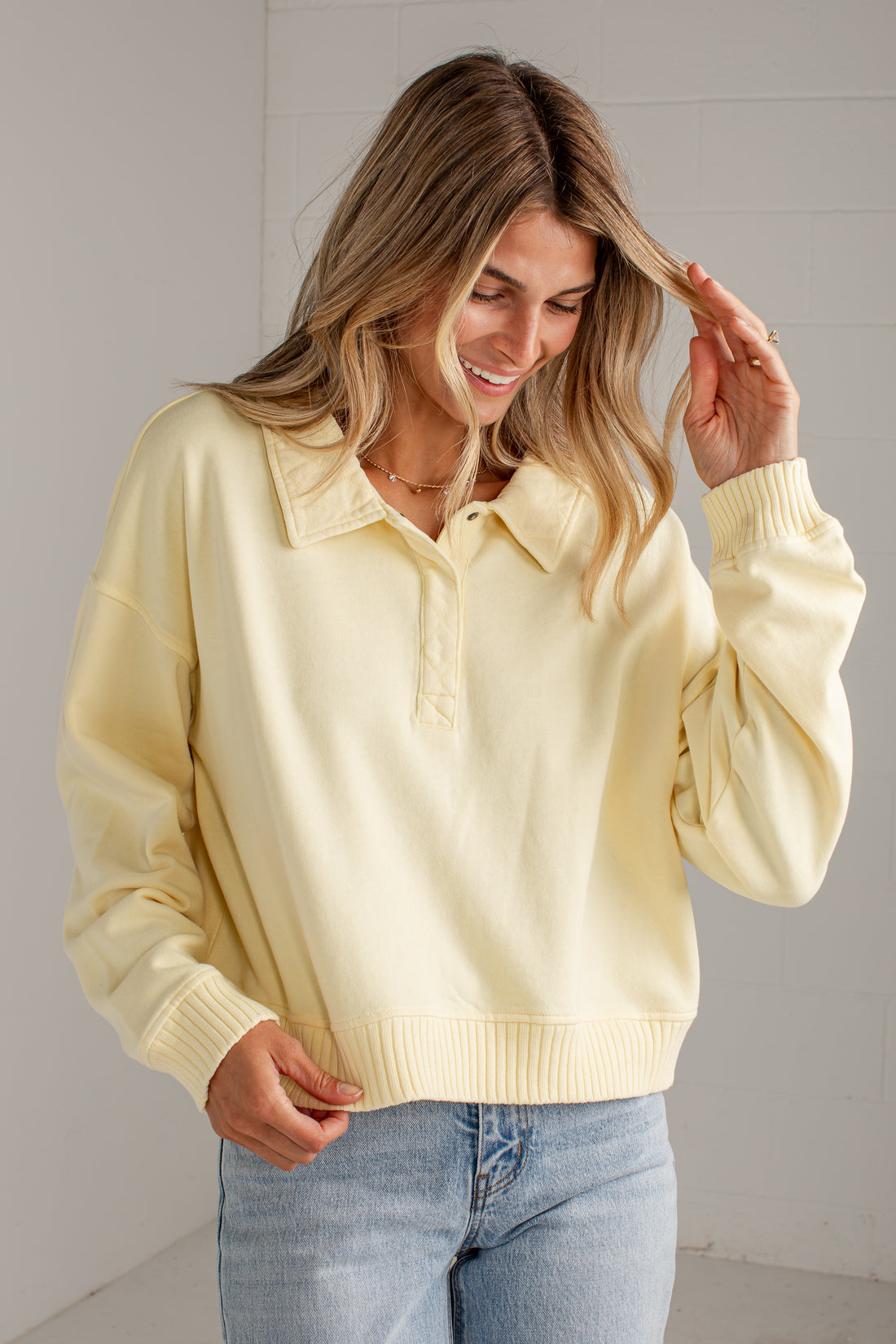 A woman wearing a pale yellow long-sleeve knit polo-style sweater with a ribbed collar, button placket, and cuffs, paired with light blue jeans, standing against a light gray wall.