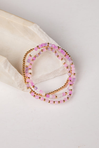 Three beaded stretch bracelets in pink and gold, displayed on a light beige textured stone prop against a white background.
