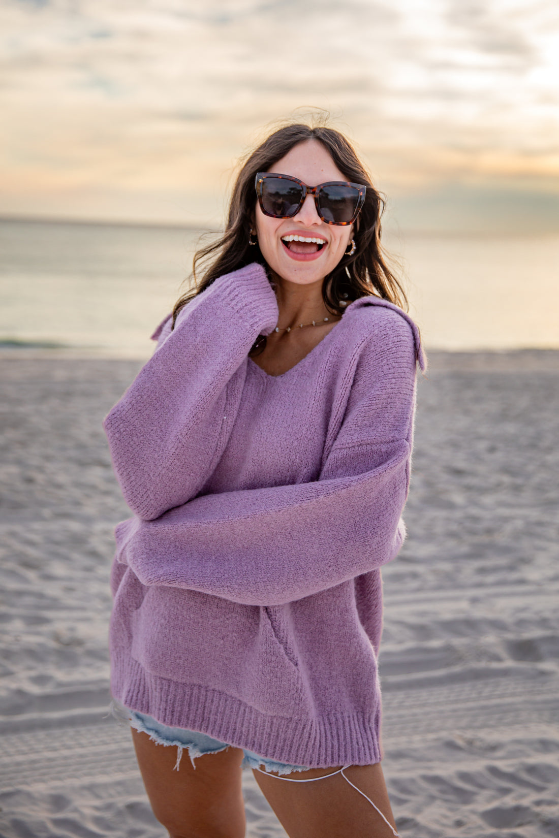 Woman wearing a purple sweater and sunglasses on a beach at sunset