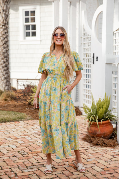Woman in a yellow floral dress standing on a brick patio with a white house in the background.