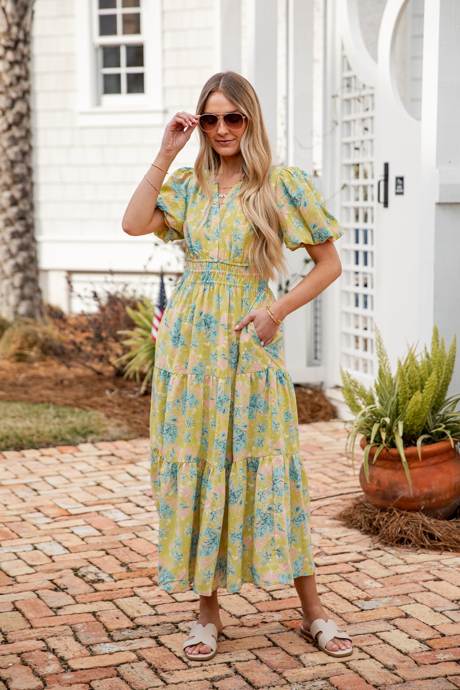 Woman in a lime floral dress standing on a brick patio with plants and a white door in the background.