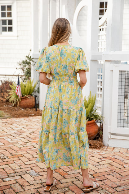 Woman wearing a yellow floral dress standing on a brick patio.