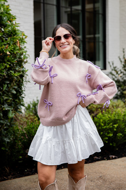 Woman wearing a pink sweater with purple bows and a white skirt outdoors.