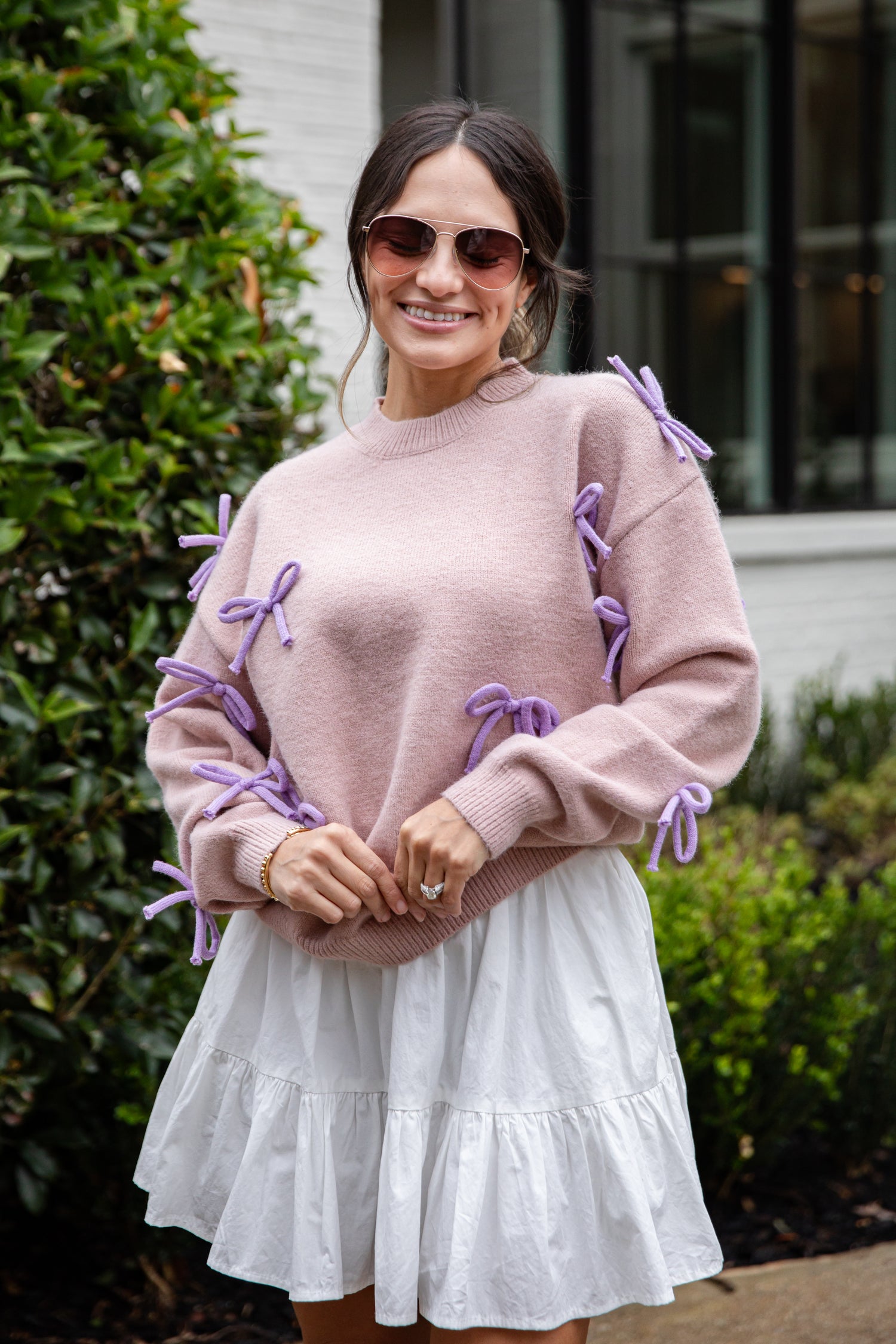 Woman wearing a pink sweater with purple accents and a white skirt outdoors.