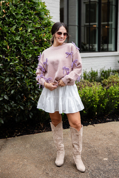 Woman wearing a pink sweater with purple bows, white skirt, and beige knee-high boots standing outdoors.