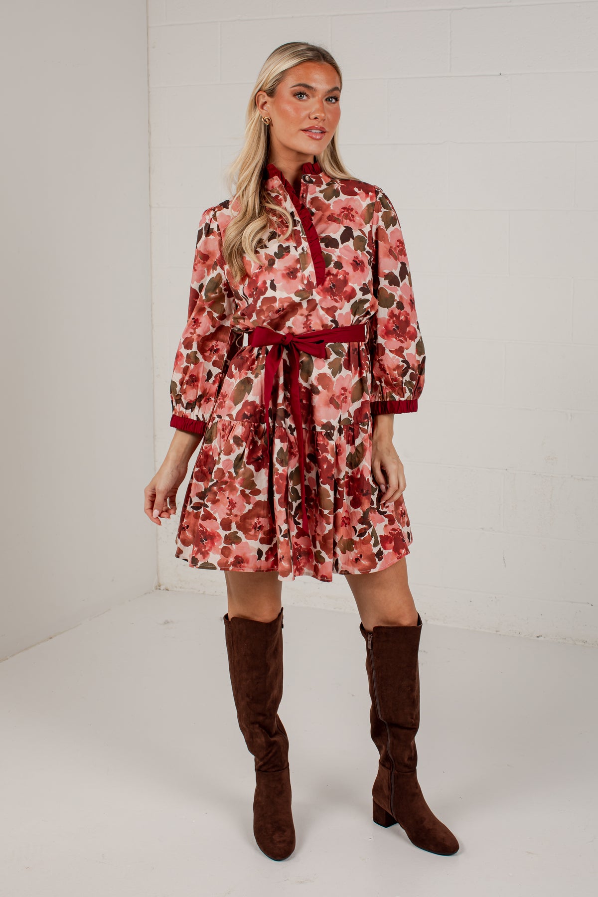 A woman models a long-sleeve floral print dress with a red tie-neck and waist belt, paired with brown knee-high boots, in a white studio setting.