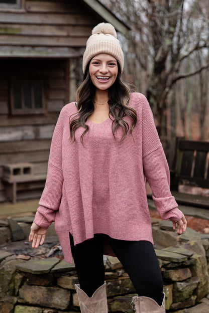 Woman wearing a pink sweater and beige beanie outdoors near a wooden cabin.