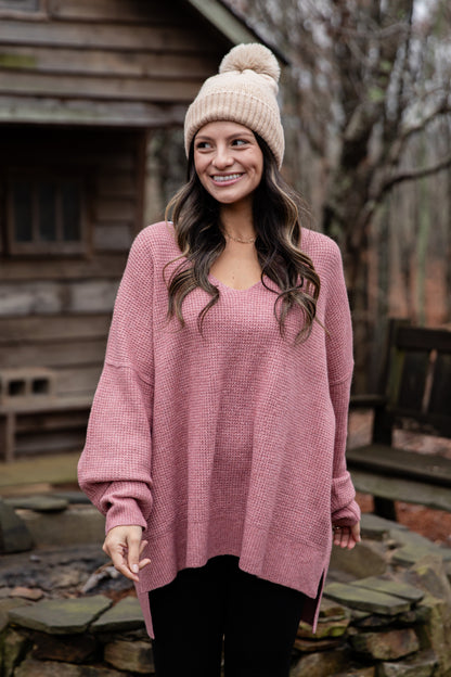 Woman wearing a pink sweater and beanie standing outdoors near a wooden cabin.