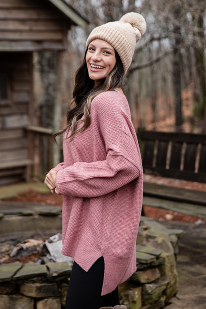 Woman wearing a pink sweater and beanie standing outdoors near a wooden cabin.