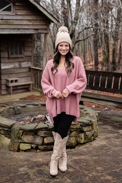 Woman in pink sweater and white boots standing in front of a wooden cabin.
