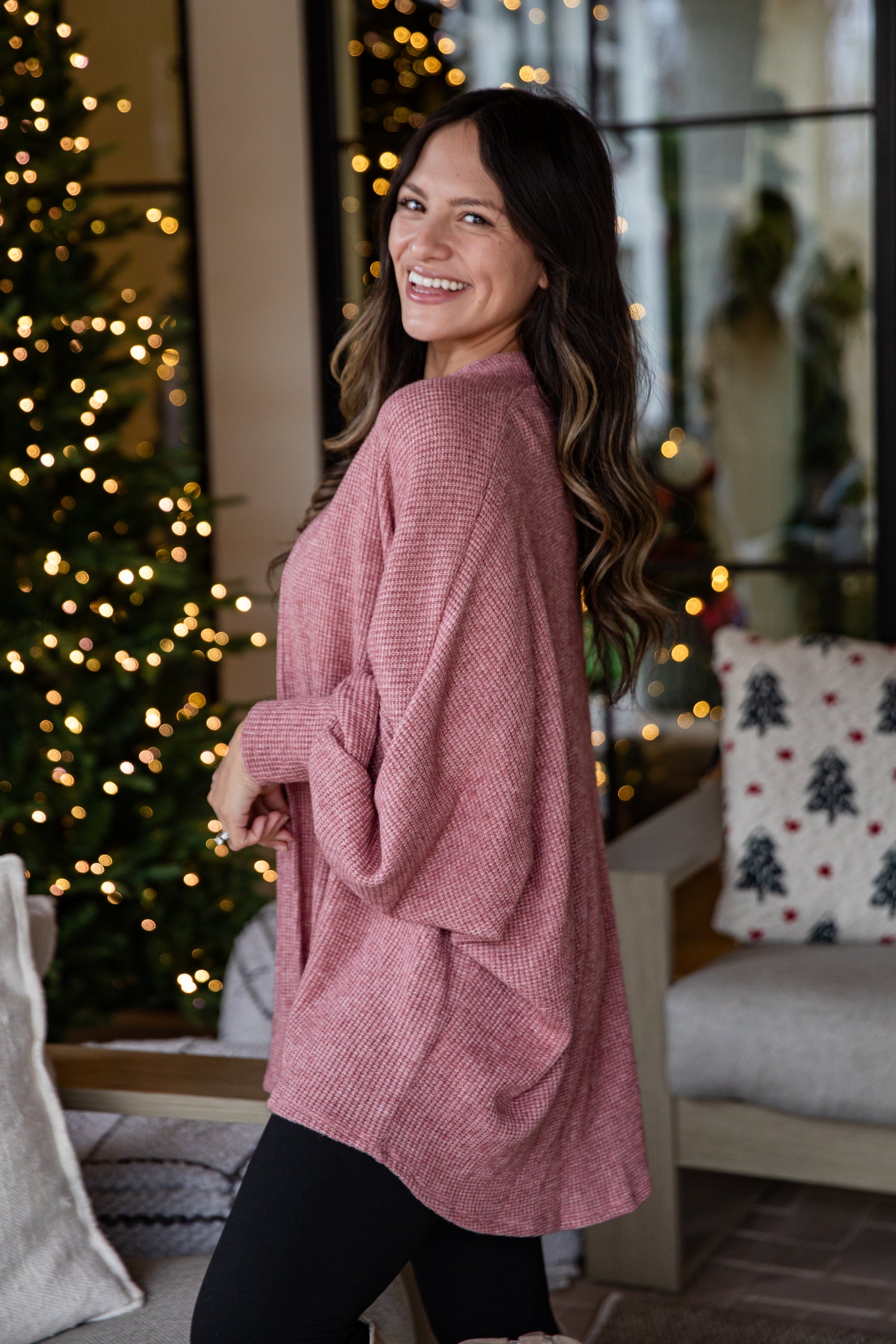 Woman wearing a pink sweater standing in a festive indoor setting with Christmas decorations.