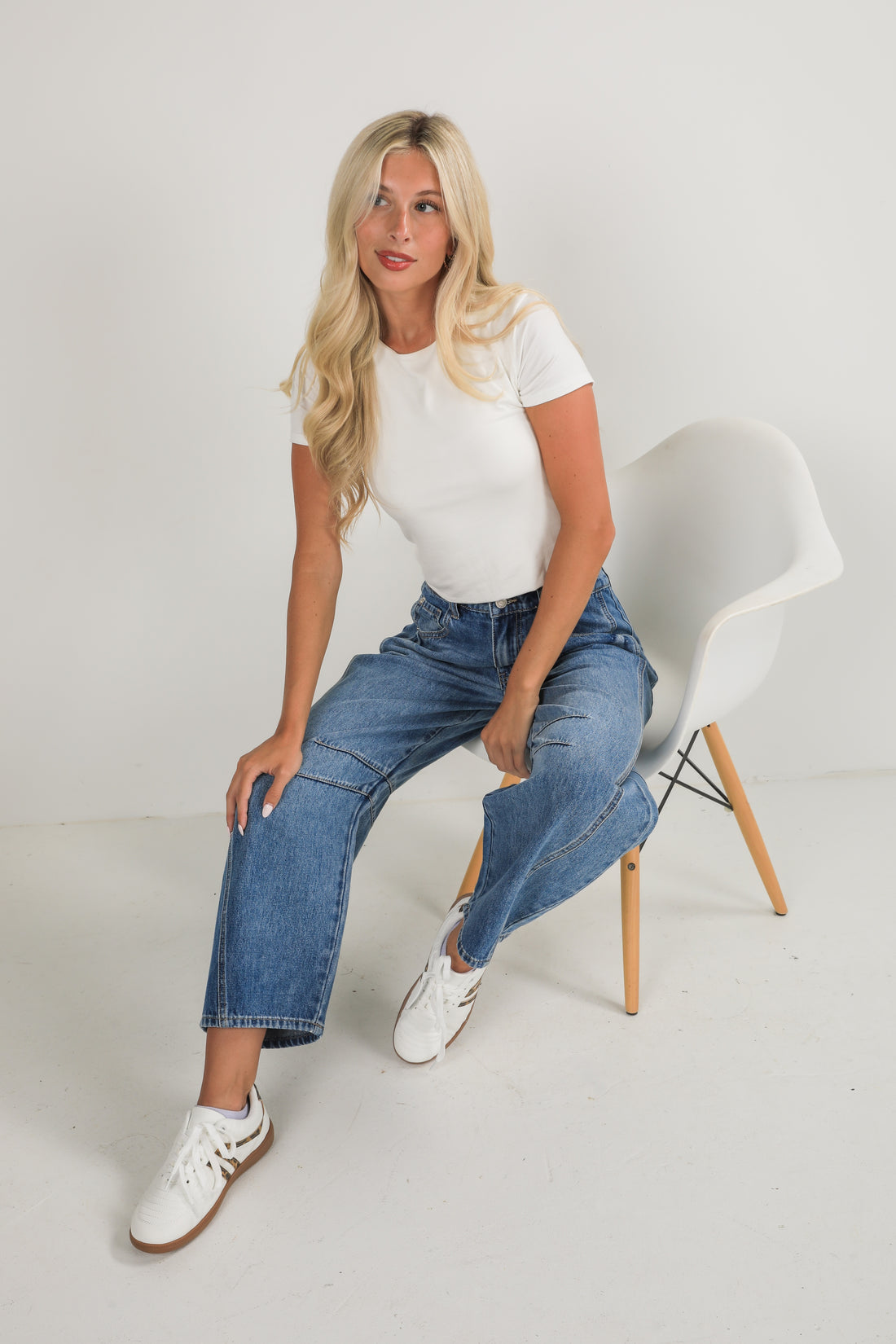 Woman sitting on a white chair wearing a white t-shirt, blue jeans, and white sneakers.