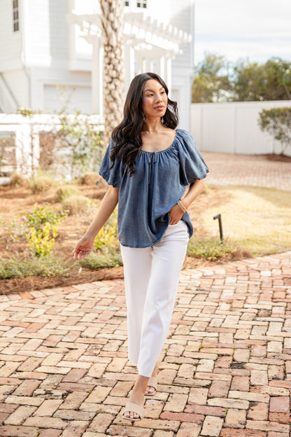 Woman in a blue top and white pants walking on a brick path outdoors.