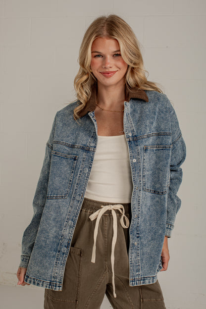 A woman wearing a light blue acid-wash denim shirt jacket with a brown collar, styled over a white tank top and olive green drawstring cargo pants, standing against a light gray brick wall.