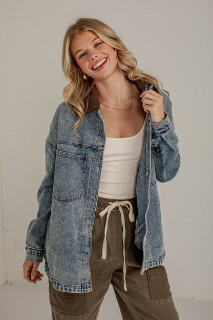 A woman smiling and wearing an oversized blue denim jacket over a white tank top and olive green drawstring pants, standing against a light gray wall.